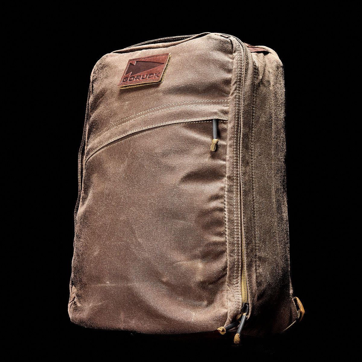 A brown GR1 USA - Heritage Waxed Canvas rucksack with a leather patch and visible zippers is shown on a black background, crafted from durable waxed duck canvas.