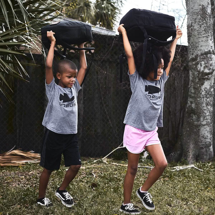 Two children outdoors lift backpacks over their heads, showcasing movements from the GORUCK Elementary School Ruck Training Program. They are dressed in matching gray t-shirts and black athletic shorts paired with sneakers. In the background, a chain-link fence and trees suggest they are in a park—the ideal setting for this training program.