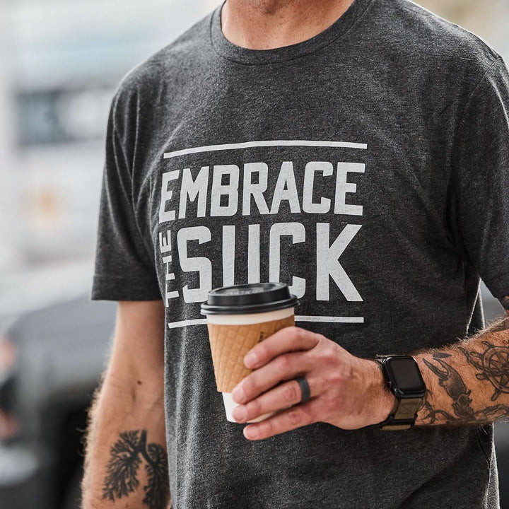A person holds a coffee cup while wearing the Embrace the Suck Tee - Tri-Blend, featuring the USMC-inspired motto "Embrace The Suck"—a reminder to face any challenge head-on.