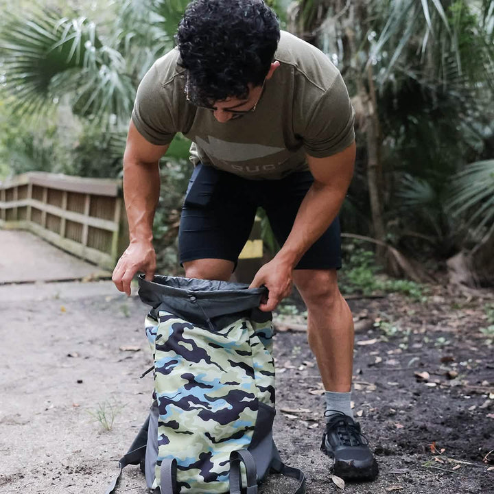 A person with short curly hair is outside, peering into an open M24 - Cordura rucksack by GORUCK. They're dressed in a green T-shirt, black shorts, and sturdy hiking shoes. In the background, you can see a wooden pathway and lush green foliage.
