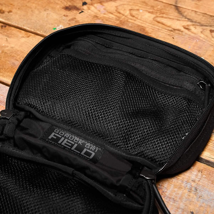 Place the black backpack featuring a mesh pocket on a wooden surface; it serves as an ideal travel accessory. The label displays "GORUCK GR1 Field Pocket - Dyneema," designed with durability for adventure seekers in mind.
