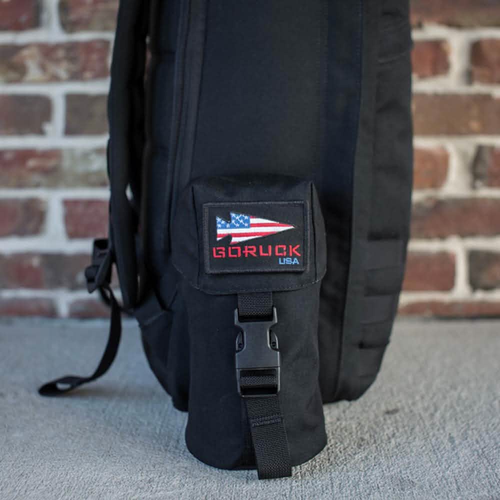 A black backpack with a Full Panel Water Bottle Pocket, adorned with a small pouch that displays an American flag patch and "GORUCK USA" in red, is leaning against a brick wall. The design features a MOLLE attachment system and includes a pocket suitable for Nalgene-style water bottles.