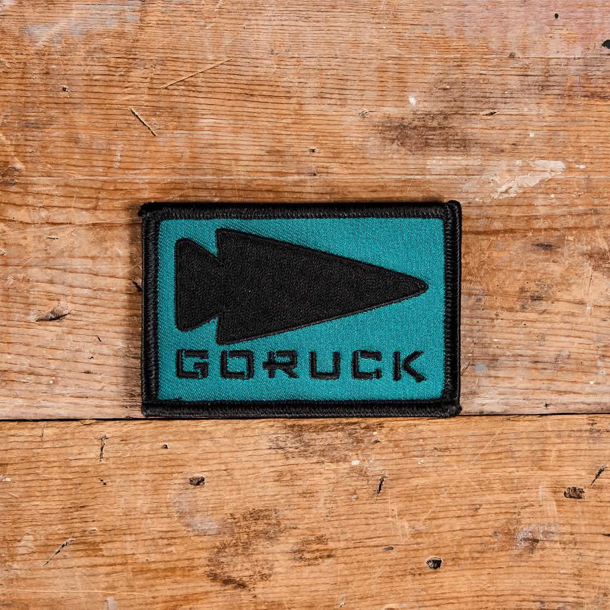 The Patch - GORUCK Spearhead by GORUCK displays a rectangular patch with a teal background, featuring an embroidered black spearhead design and the bold black GORUCK logo. This piece, backed with VELCRO, is elegantly positioned on a wooden surface.