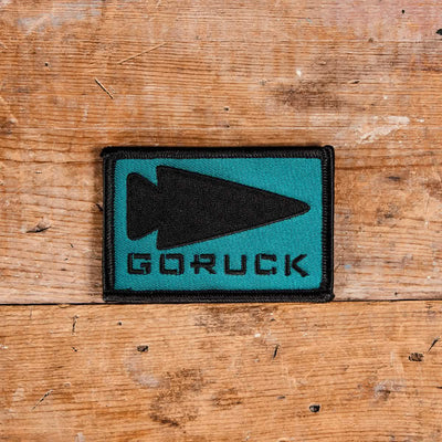 The Patch - GORUCK Spearhead by GORUCK displays a rectangular patch with a teal background, featuring an embroidered black spearhead design and the bold black GORUCK logo. This piece, backed with VELCRO, is elegantly positioned on a wooden surface.