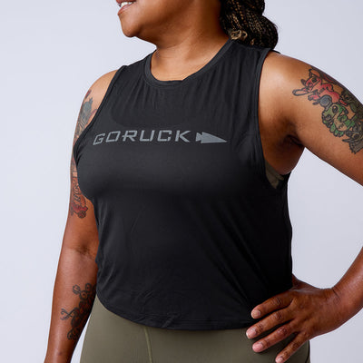 A smiling, tattooed woman wears a black Women’s USA Performance Tank - ToughMesh, featuring featherweight ToughMesh fabric, and olive leggings with a hand on her hip.