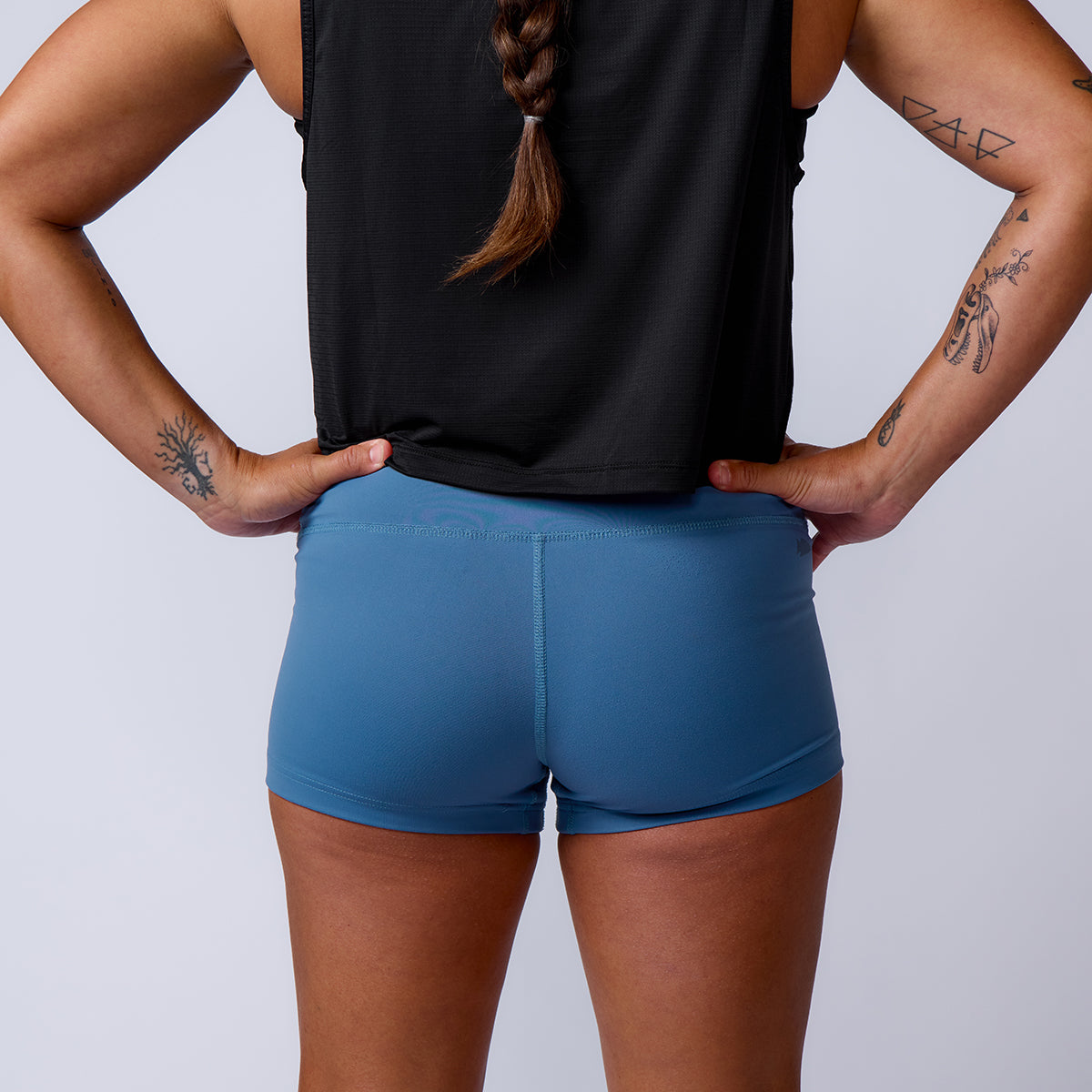 Person with tattoos wearing a black top and high-waisted blue Women’s Squat Shorts - ToughFlex, standing with hands on hips, facing away.