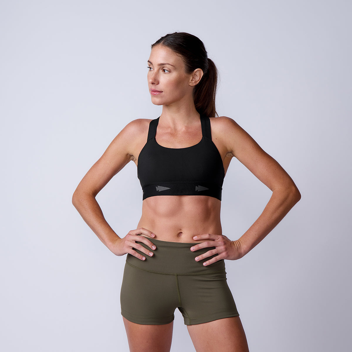 A woman in a black sports bra and high-waisted olive green Women’s Squat Shorts - ToughFlex stands with hands on hips, looking to the side.