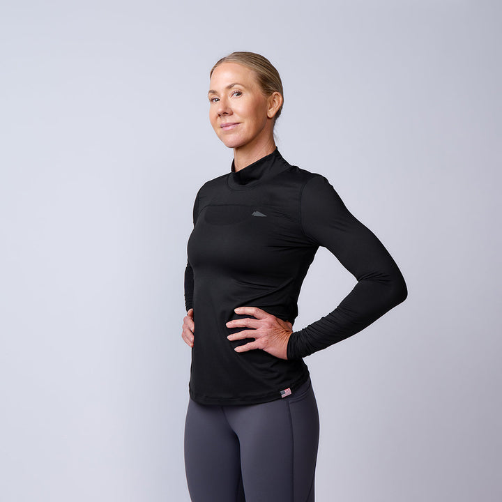 A confident woman wears the Women’s Long Sleeve Mock Neck - ToughMesh, a lightweight base layer crafted with Italian ToughMesh for enhanced breathability, set against a plain light background.