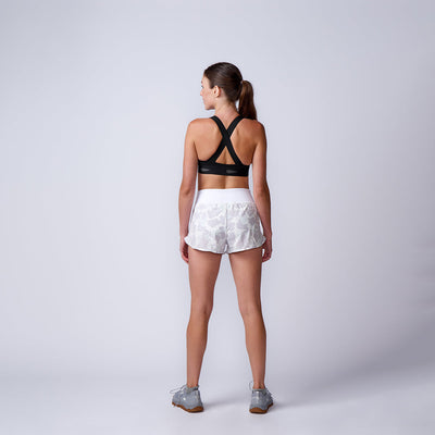 A woman stands with her back to the camera, wearing a black sports bra, Women’s Training Shorts - ToughStretch, and gray sneakers against a plain background.