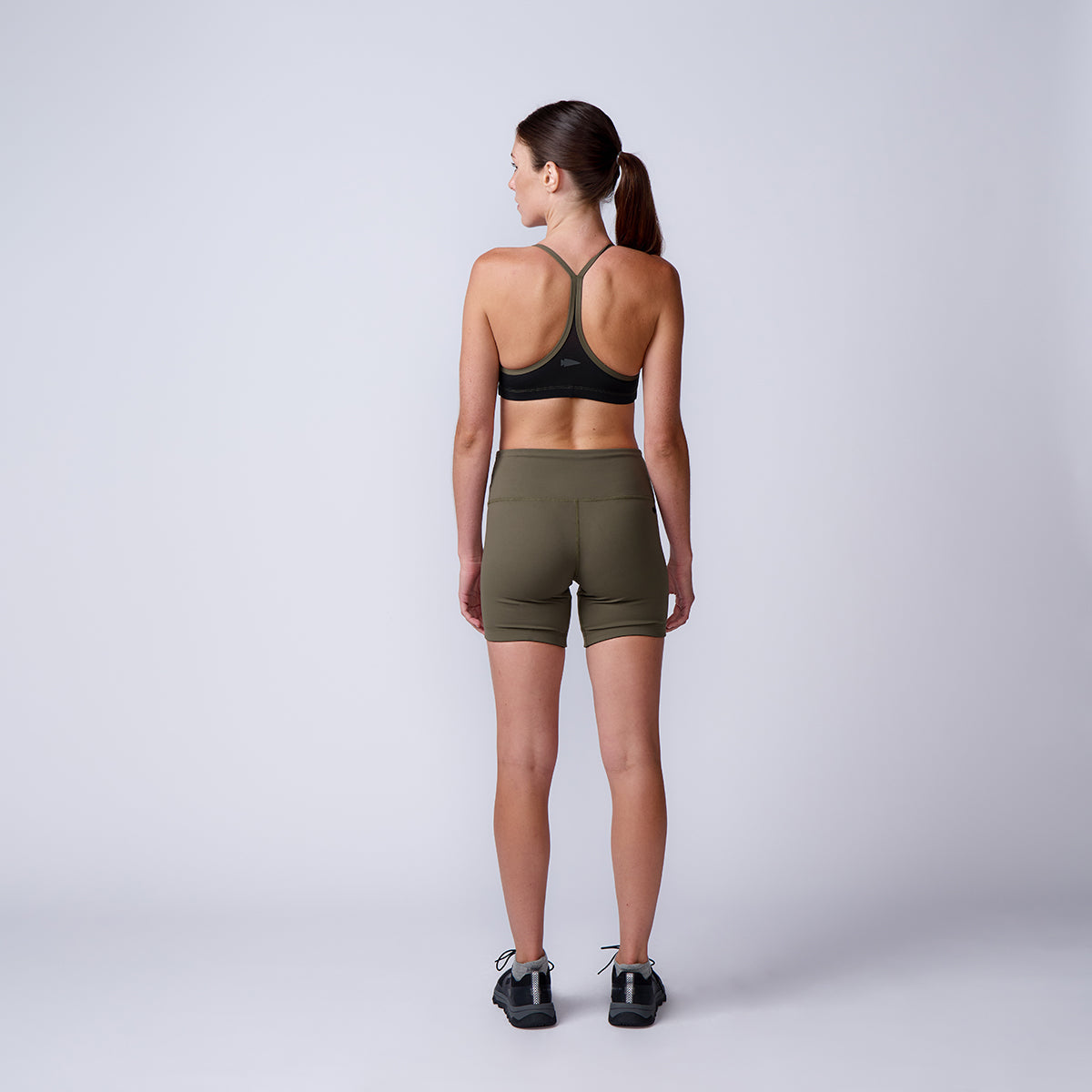 A woman stands facing away in activewear, wearing a sports bra, sneakers, and Women’s Biker Shorts - ToughFlex against a plain background.