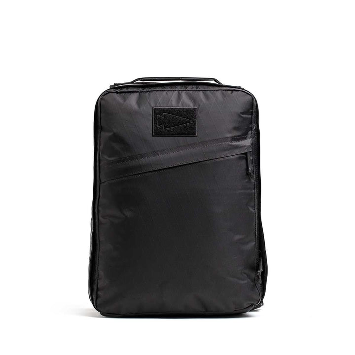 The GR0 - X-PAC is a black rectangular backpack with a top handle and diagonal front zipper pocket, made from waterproof material, displayed against a white background.