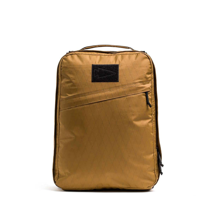 The GR0 - X-PAC is a tan, rectangular, lightweight rucksack made from waterproof material, featuring a front zipper pocket and a black patch logo.
