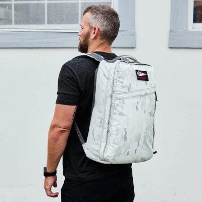 A man with a beard stands sideways, wearing a black T-shirt and the GR1 USA - Cordura (The Original Ruck) backpack by GORUCK, which features a light gray camouflage design suggestive of Special Forces precision. He is outside, in front of an impressive American-made structure—a light-colored building with two windows framed in gray.