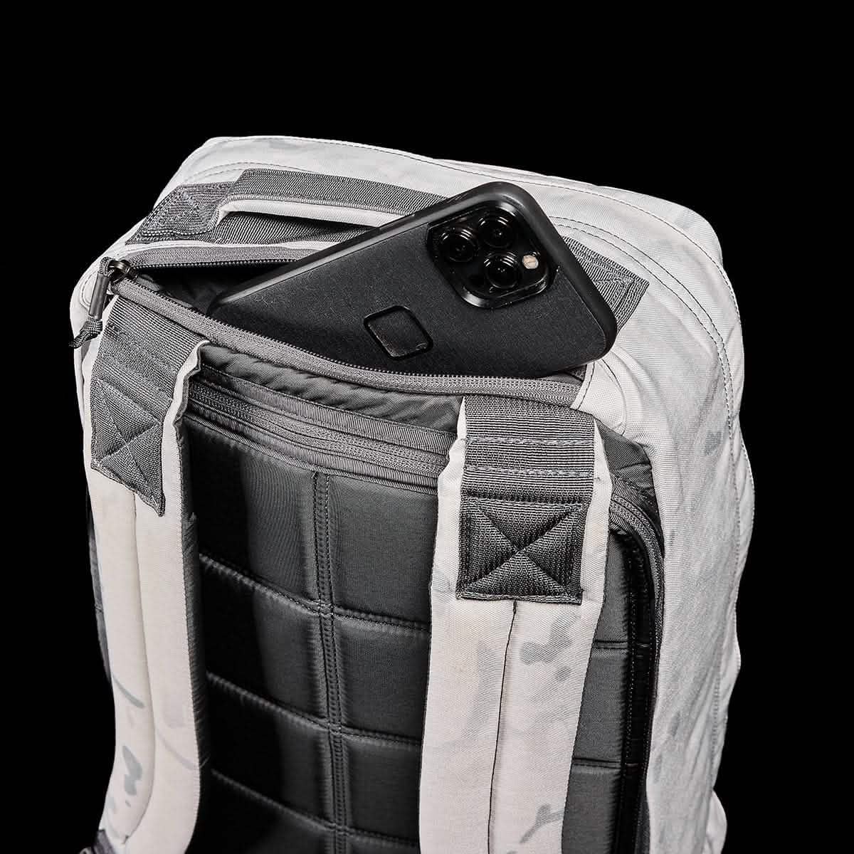 A sleek black smartphone peeks from the back zipper pocket of the GR1 USA - Cordura (The Original Ruck) backpack from GORUCK, its gray and black padding exuding durability. Set against a solid black backdrop, this sturdy pack is ready for any GORUCK Challenge, highlighting its quilted texture and Special Forces-inspired design.