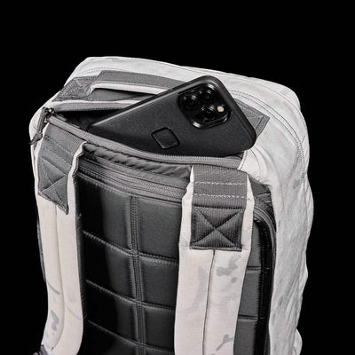 A sleek black smartphone peeks from the back zipper pocket of the GR1 USA - Cordura (The Original Ruck) backpack from GORUCK, its gray and black padding exuding durability. Set against a solid black backdrop, this sturdy pack is ready for any GORUCK Challenge, highlighting its quilted texture and Special Forces-inspired design.
