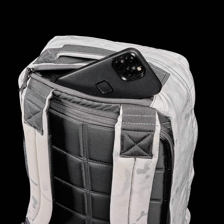A sleek black smartphone peeks from the back zipper pocket of the GR1 USA - Cordura (The Original Ruck) backpack from GORUCK, its gray and black padding exuding durability. Set against a solid black backdrop, this sturdy pack is ready for any GORUCK Challenge, highlighting its quilted texture and Special Forces-inspired design.