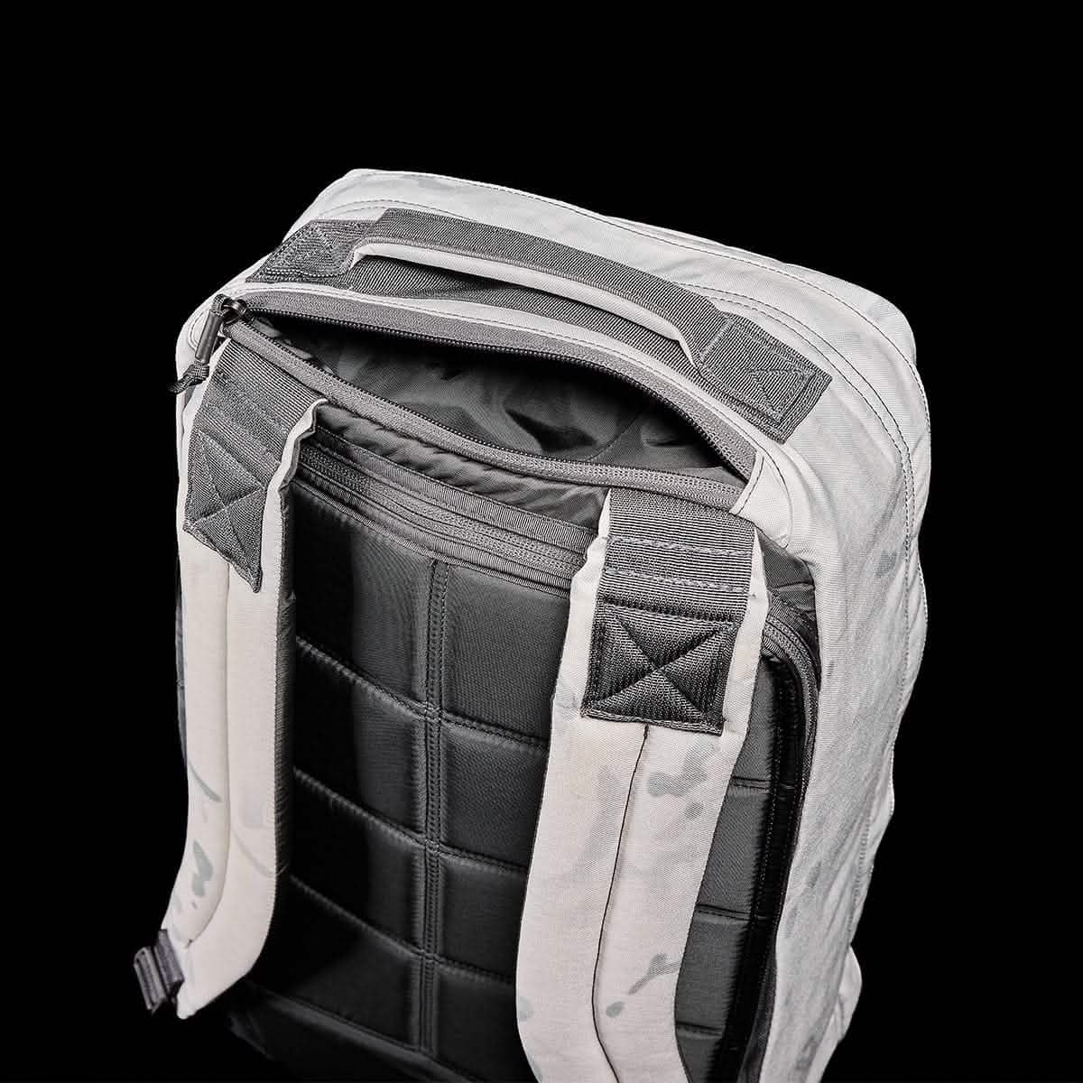 A GR1 USA backpack from GORUCK, made of Cordura, is shown against a black background. It features gray fabric with black padded straps and a quilted back panel. The top zipper is slightly open, exposing the interior lining. Designed to endure the challenges of a GORUCK event, the reinforced handle at the top ensures durability.