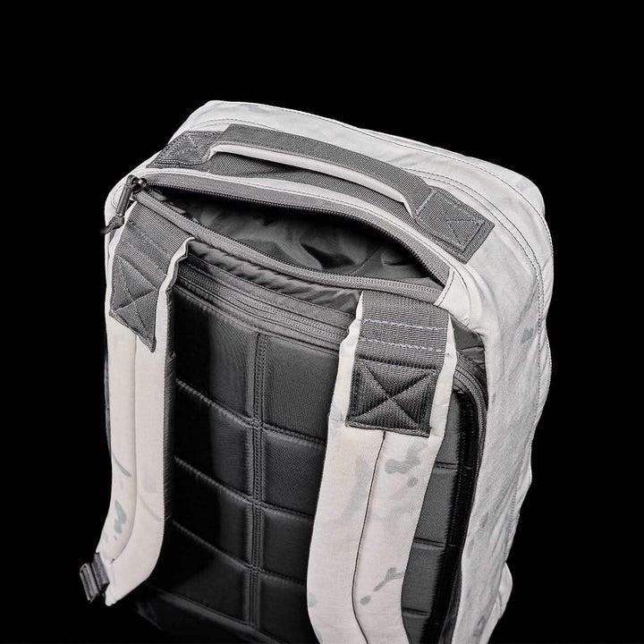 A GR1 USA backpack from GORUCK, made of Cordura, is shown against a black background. It features gray fabric with black padded straps and a quilted back panel. The top zipper is slightly open, exposing the interior lining. Designed to endure the challenges of a GORUCK event, the reinforced handle at the top ensures durability.