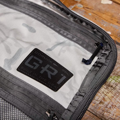Zoom in on the interior label of the GR1 USA - Cordura (The Original Ruck) by GORUCK, showcasing its "Built in the USA" heritage, inspired by Special Forces. The gray and black design is highlighted by a prominent zipper and precise stitching, as the backpack sits on a wooden surface, poised for any GORUCK Challenge.