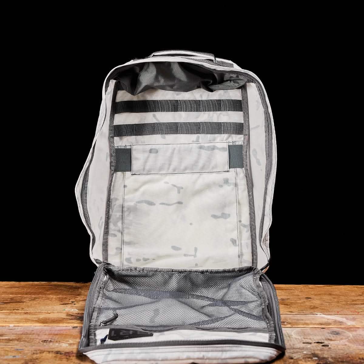 The GR1 USA - Cordura (The Original Ruck) by GORUCK is displayed open, revealing its spacious main compartment. This gray backpack captures the rugged essence and American manufacturing quality reminiscent of the GR1 design. It features two horizontal black straps and a large internal pocket, all set against a wooden surface with a black background.
