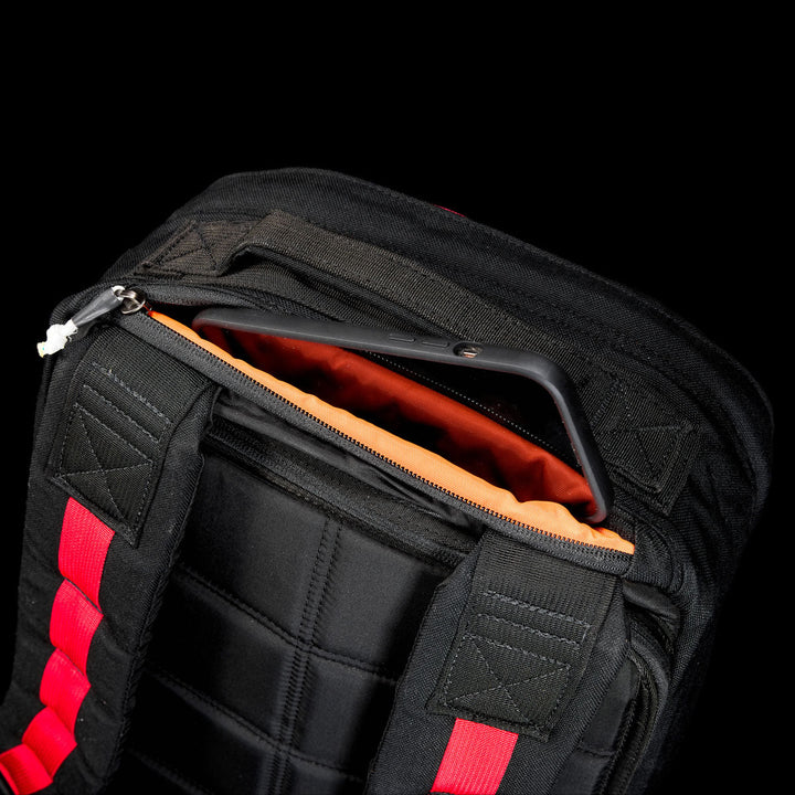 The GR1 Shooter 26L - Built by Scars backpack features a durable CORDURA® exterior, red straps, an open compartment with orange lining, and fits a black smartphone inside.