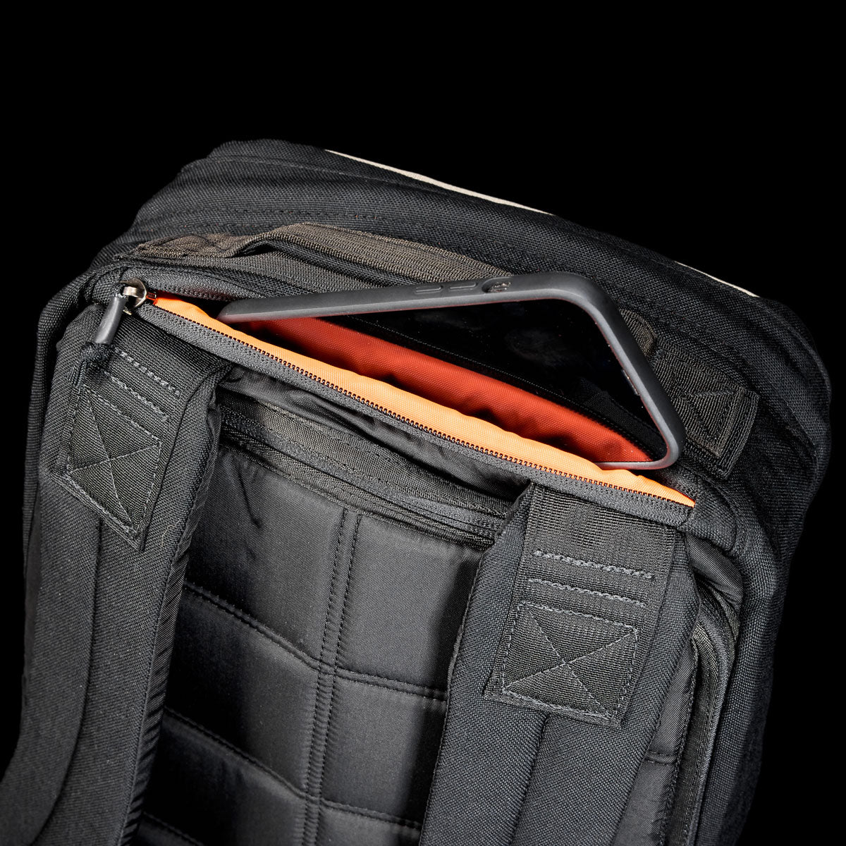A tablet partially slides out of an orange-lined pocket in the black GR1 Shooter 26L - Built by scars backpack, made from durable CORDURA®, set against a black background.