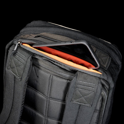 A tablet partially slides out of an orange-lined pocket in the black GR1 Shooter 26L - Built by scars backpack, made from durable CORDURA®, set against a black background.