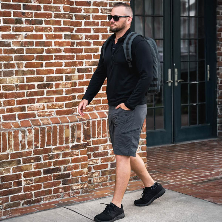 A man wearing sunglasses, a black long-sleeve shirt, gray shorts, and black shoes walks along a sidewalk. Sporting the rugged GR1 USA - Cordura (The Original Ruck) by GORUCK, he strides confidently in front of a brick wall with glass doors.
