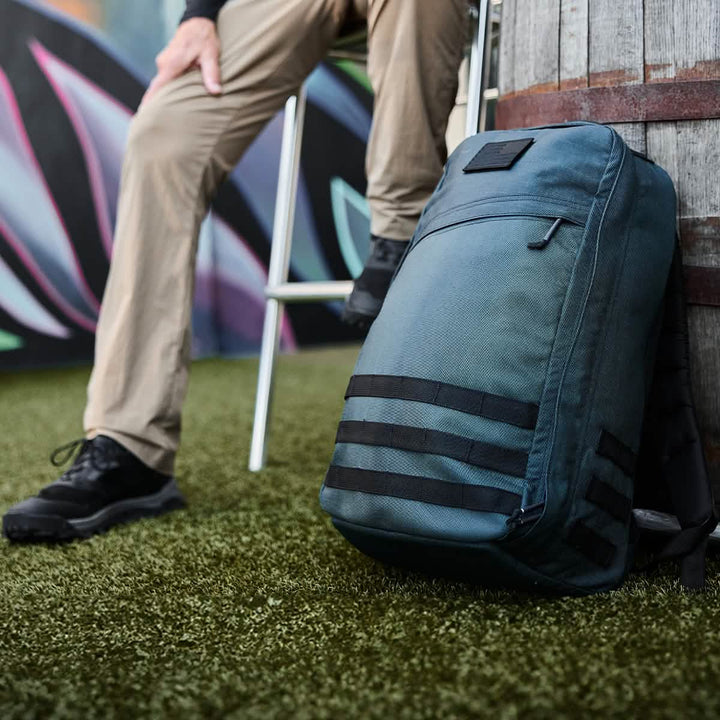 A person sits on a stool on artificial grass, clad in khaki pants and black shoes that evoke the rugged style of a Special Forces operative. Beside them rests a GR1 USA - Cordura (The Original Ruck) backpack by GORUCK, featuring distinctive black horizontal straps as it leans against a barrel. In the background, vibrant graffiti adds a splash of color.