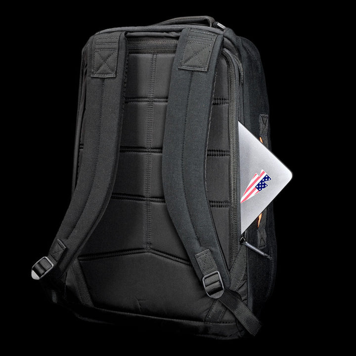 The GR1 Shooter 26L - Built by Scars backpack, crafted from Special Forces Grade CORDURA®, with visible straps, holds a laptop featuring a US flag sticker in the side pocket, all set against a black background.