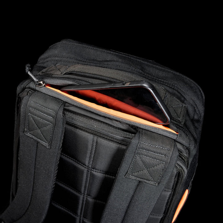 A smartphone partially tucked in a zippered pocket on a black GR1 Shooter 26L - Built by Scars backpack, crafted from Special Forces Grade CORDURA® with padded straps.