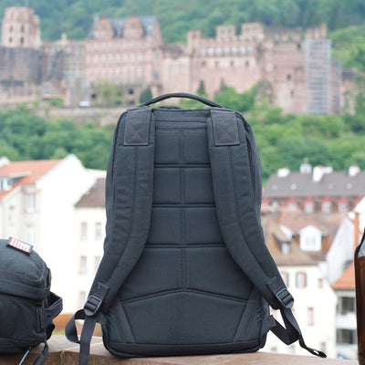 A black GR1 USA Slick - Ballistic Nylon Cordura backpack stands upright outdoors, showcasing its bombproof laptop compartment with a blurred castle and buildings in the background.