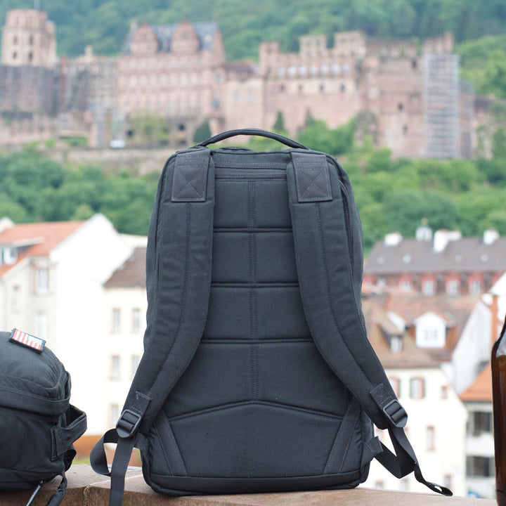 A black GR1 USA Slick - Ballistic Nylon Cordura backpack stands upright outdoors, showcasing its bombproof laptop compartment with a blurred castle and buildings in the background.
