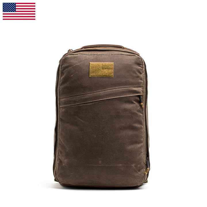 The GR1 USA - Heritage Waxed Canvas is a durable backpack made from waxed duck canvas, featuring a tan patch and a small American flag in the top left corner.