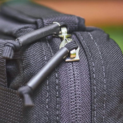 A close-up image of the GR1 Field Pocket, a black electronics organizer by GORUCK, with a silver zipper partially open. It showcases thick black stitching and sturdy zipper pulls. The blurred background highlights the texture and details of the organizer's material.