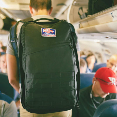 A person wearing a large black GR2 by GORUCK is standing in the aisle of an airplane. The bag features a patch with a Special Forces logo. In the background, passengers are seated, with one individual wearing headphones and a red cap.