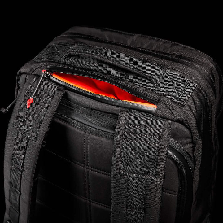 A close-up of the GR2 - Ripstop ROBIC® backpack by GORUCK showcases its black exterior with padded straps and a partially open top zipper pocket, revealing a bright orange interior. The textured, water-resistant fabric and stitching details of the rucksack stand out against a dark background.