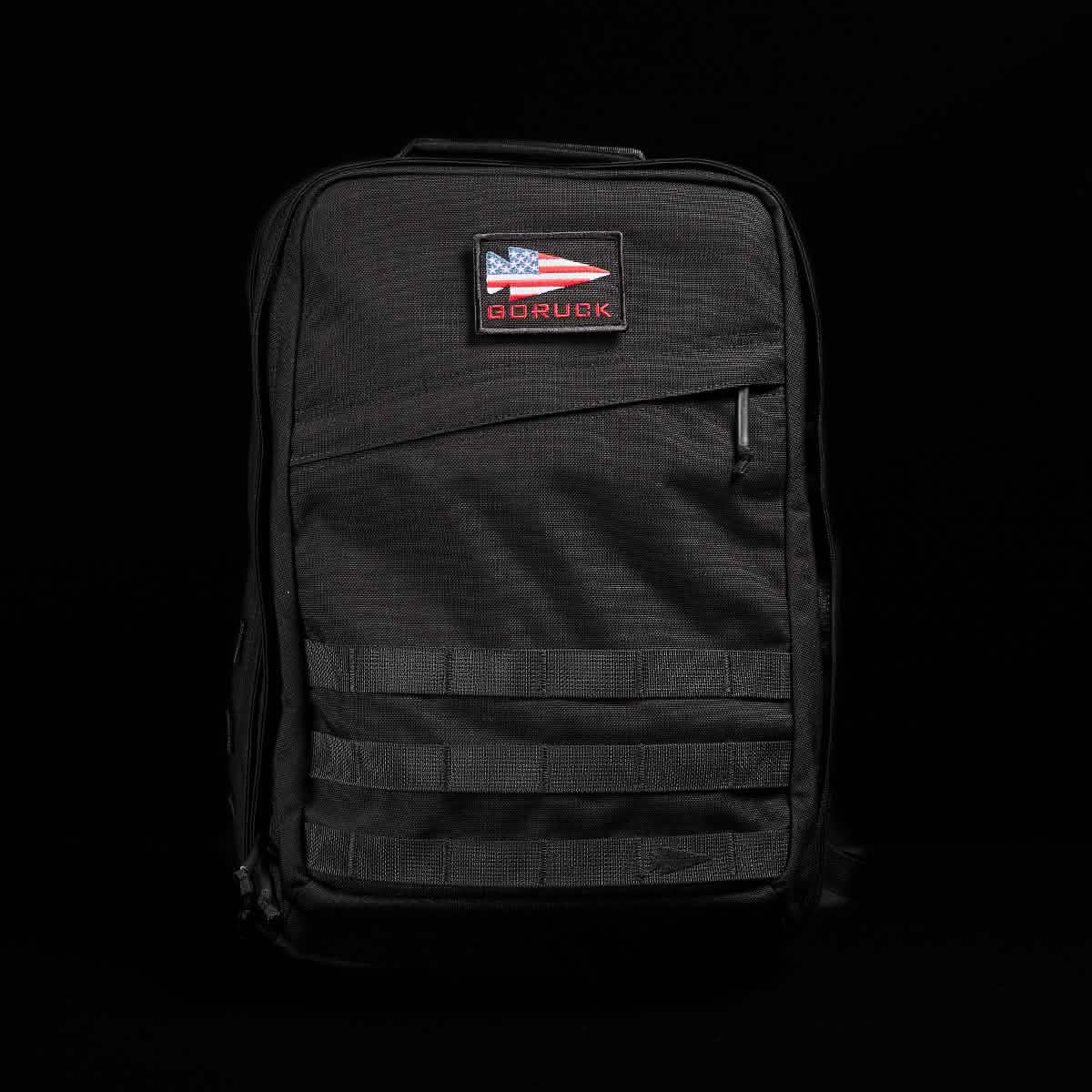 A black GR2 - Cordura backpack from GORUCK, reminiscent of Special Forces gear, is displayed against a dark background. It features a patch with an American flag and the brand name. The ruck boasts multiple compartments and a sleek, rugged design.