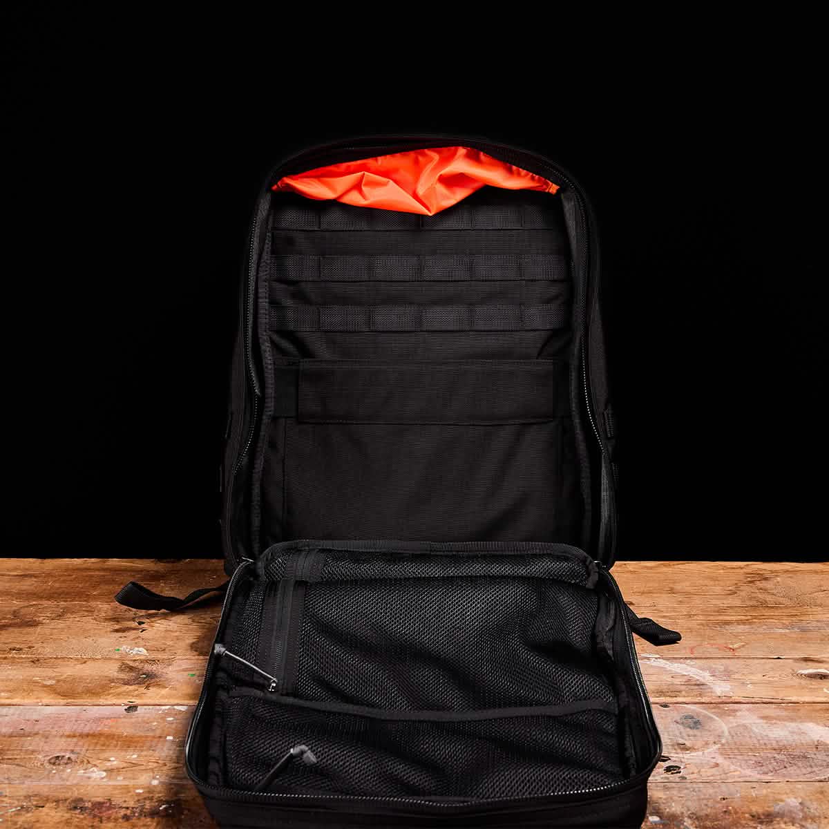 The GR2 - Cordura by GORUCK, an open black backpack echoing the design of a Special Forces ruck, sits on a wooden surface. Inside, its empty space reveals a bright orange lining at the top along with a mesh pocket and several organizational slots set against the stark black background.