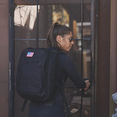 A woman with long hair, dressed in sunglasses and a black outfit, carefully maneuvers a lock as she quietly opens a gate. Her GORUCK GR2 - Cordura backpack features an American flag patch. The scene is outdoors near a building, highlighting the precision of Special Forces in her assured actions.