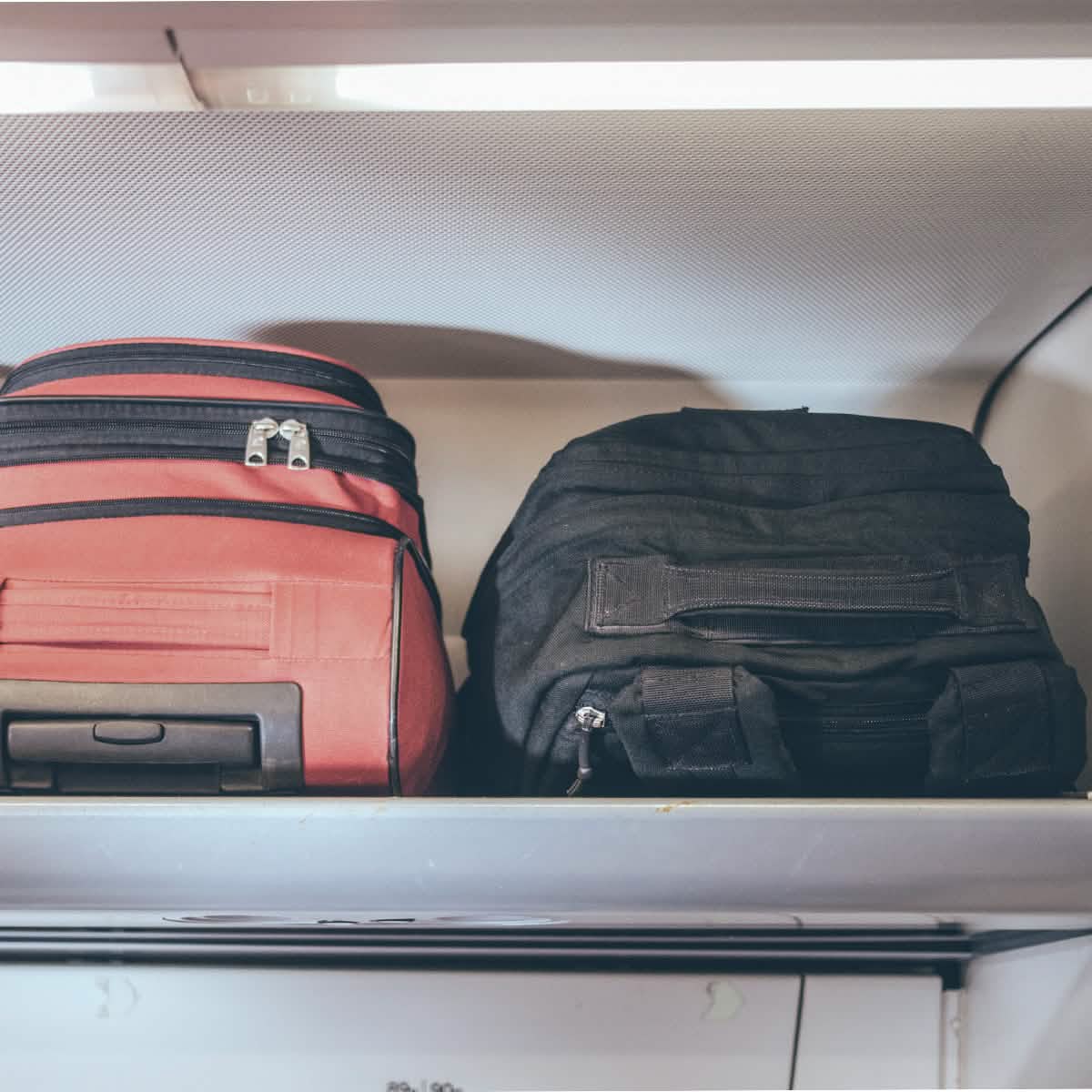 A red GR2 suitcase and a black GORUCK travel bag are stored in an airplane's overhead compartment.