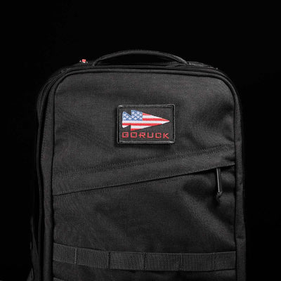 Set against a dark backdrop, the stylish GR2 backpack by GORUCK features a patch with an American flag and 'GORUCK' in red. Designed with Special Forces quality and a robust top handle, it ensures perfect carry-on compliance.