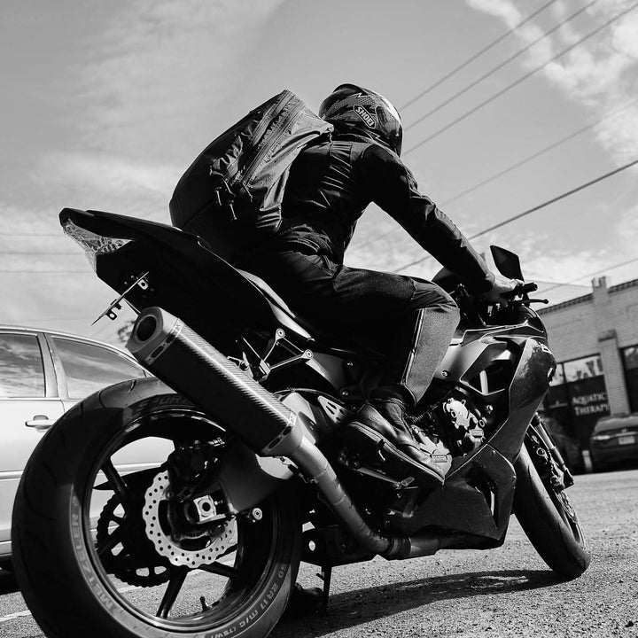 A motorcyclist wearing a helmet and a GR2 X-PAC backpack with AquaGuard zippers rides a sport bike on a city street during the day.