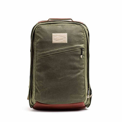 The GR2 - Waxed Canvas is an olive green backpack with a brown leather base, front zipper pocket, and beige patch. Made from durable duck canvas, it's perfect for daily use or travel and built for lasting style.