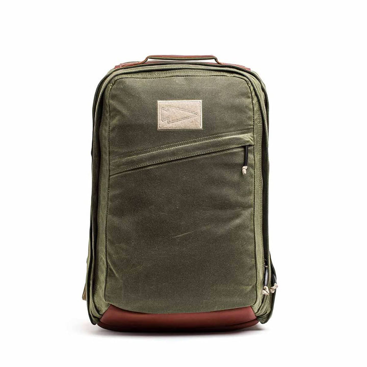 The GR2 - Waxed Canvas is an olive green backpack with a brown leather base, front zipper pocket, and beige patch. Made from durable duck canvas, it's perfect for daily use or travel and built for lasting style.
