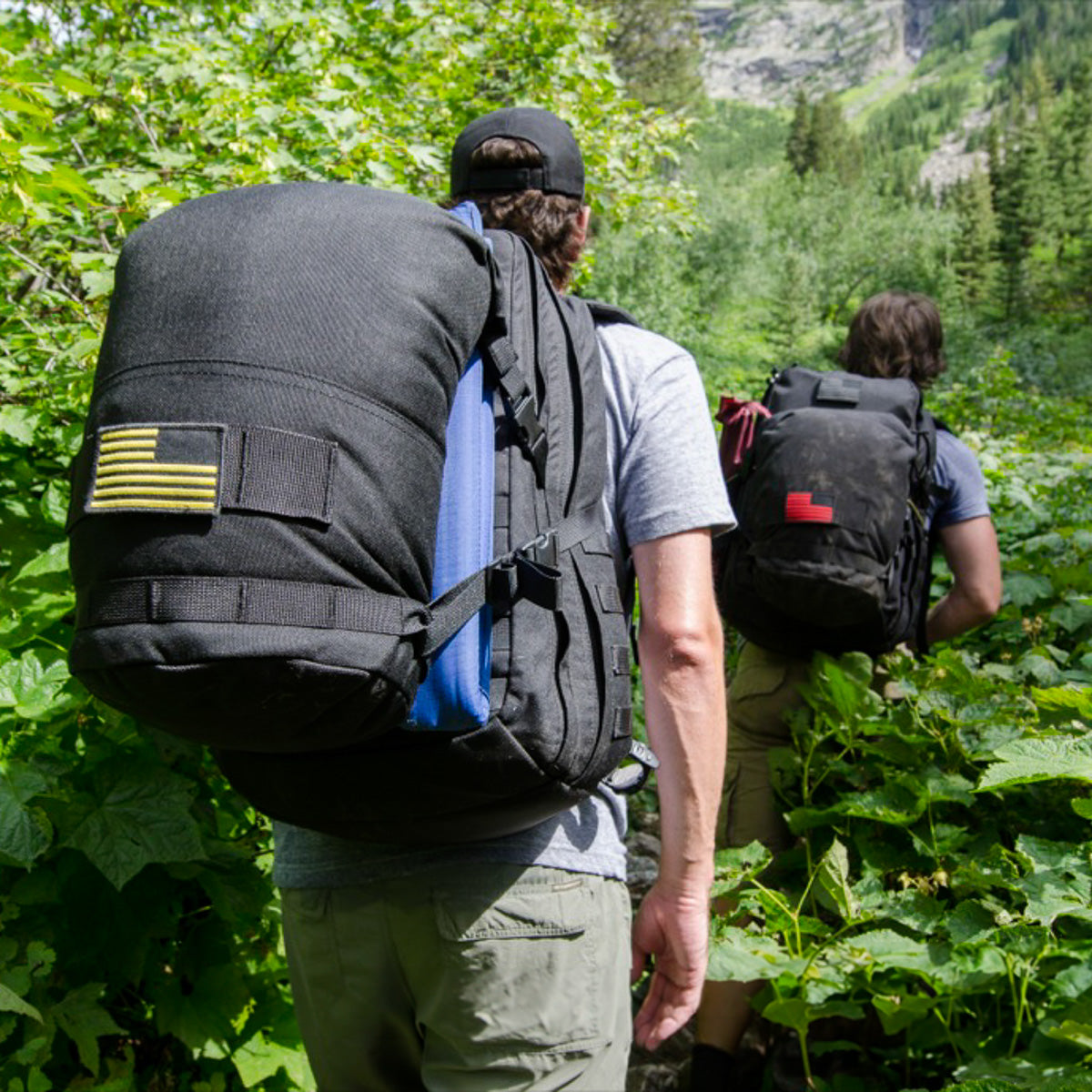 Two people with large backpacks, including the GR2 USA Throwback made from rugged 1000D Cordura, hike through lush, green vegetation on a forest trail.