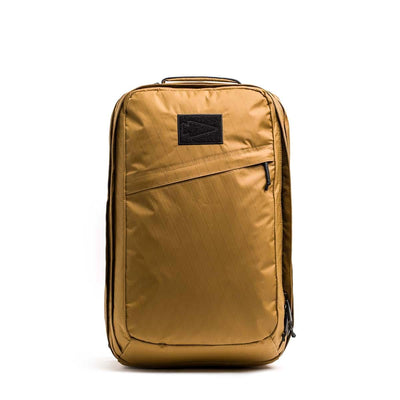 The GR2 X-PAC is a golden brown backpack featuring a front diagonal AquaGuard zipper pocket, black patch, top handle, and crafted from durable X-PAC material, shown on a white background.