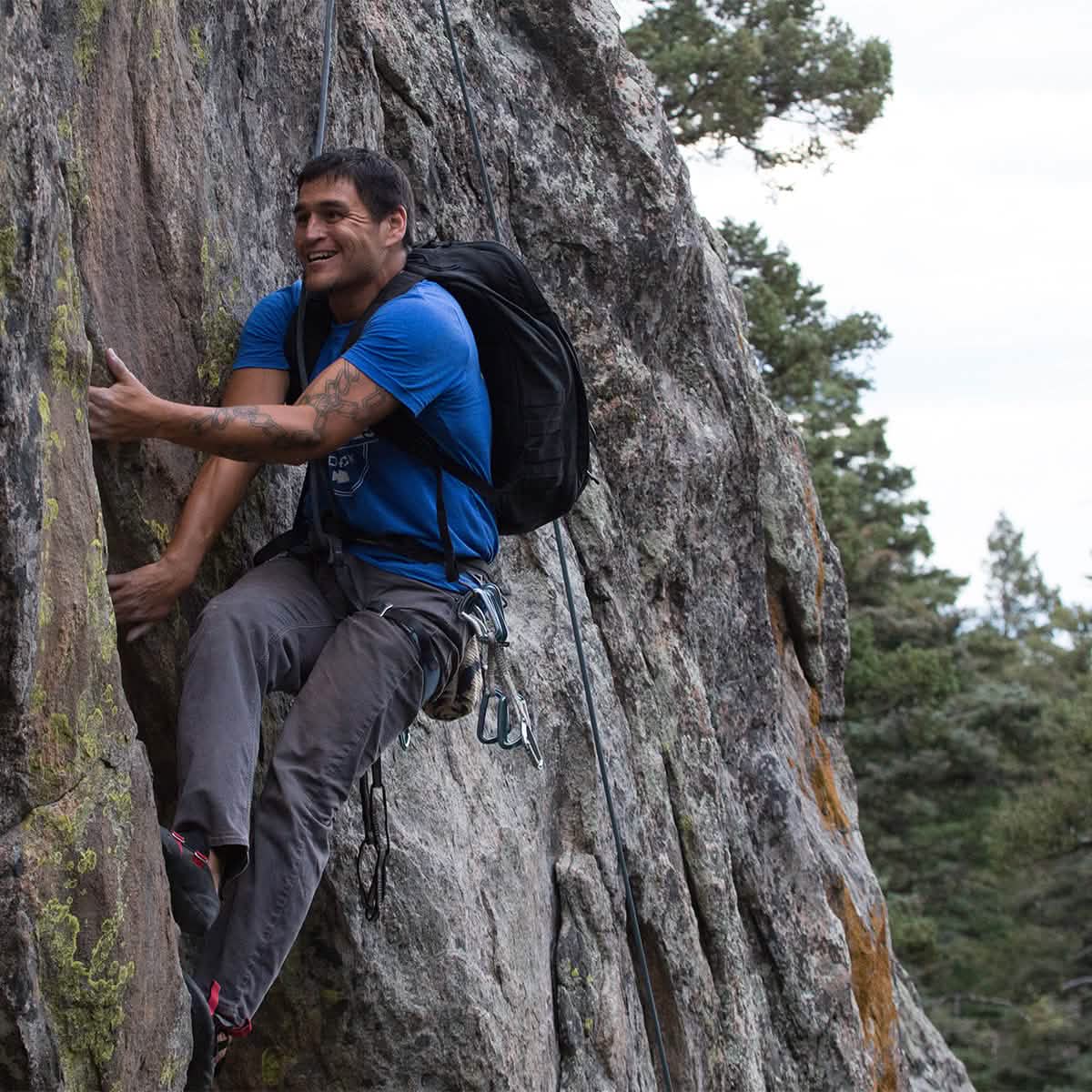 Clad in his blue shirt and climbing gear, a man with the precision reminiscent of GORUCK GR2 scales a rocky cliff while smiling, set against a stunning backdrop of trees and sky.