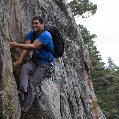 Clad in his blue shirt and climbing gear, a man with the precision reminiscent of GORUCK GR2 scales a rocky cliff while smiling, set against a stunning backdrop of trees and sky.