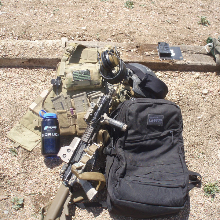 Military gear—a rifle, helmet, tactical vest, and the GR2 USA Throwback travel backpack made from tough 1000D Cordura—rests on the ground, mission-ready.